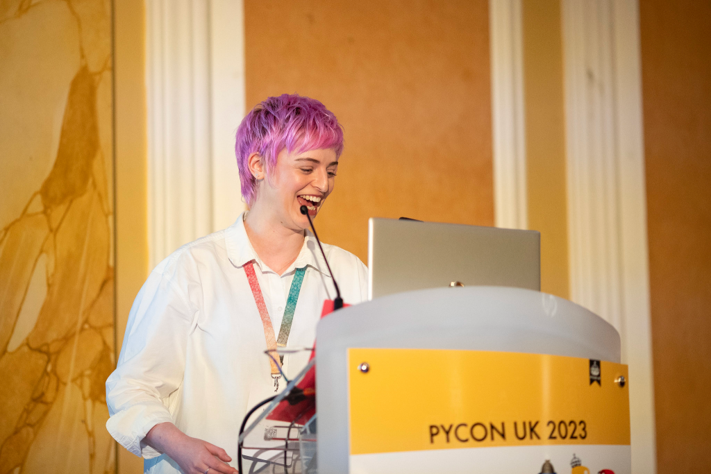 Emily, a white woman in her thirties with pink hair, speaks at a podium at PyCon UK 2023